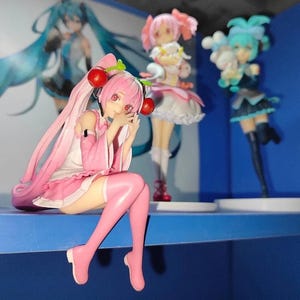 May include: A collection of anime-style figurines. The foreground figure is a pink-haired character with cherry-shaped hair accessories, wearing a pink and white outfit. Other figures in the background have different hair colors and outfits.