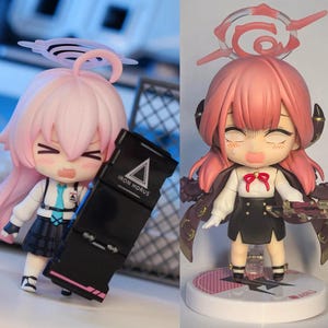May include: Two anime-style figurines with large heads and expressive faces. One figure has pink hair and holds a black object with the text "IRON HORUS". The other figure has pink hair and wears a black skirt and cape. Both figures have detailed outfits.