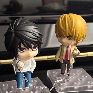 May include: Two anime-style figurines, one with black hair and the other with blonde hair, are chained together by handcuffs. The black-haired figure wears a white shirt and blue pants, while the blonde figure wears a tan jacket and red tie.