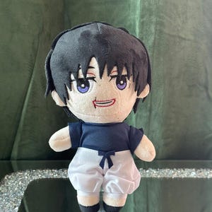 May include: A plush doll with black hair and purple eyes, wearing a navy blue shirt and white shorts. The doll has a smiling expression and black shoes. The doll is approximately 8 inches tall.