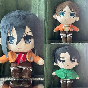 Op de afbeelding: Drie pluche poppen van anime-personages. Eén pop heeft donkerblauw haar, een oranje jas en een rode sjaal. Een andere heeft bruin haar en een oranje jas. De derde pop heeft zwart haar en een groene cape.