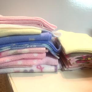 May include: A stack of soft fleece blankets in various pastel colors, including pink, blue, yellow, and white. Some blankets have patterns, such as polka dots and ribbons. The blankets are neatly folded and stacked.