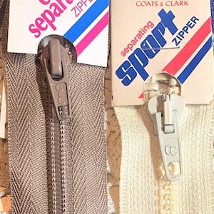 Puede incluir: Dos tiradores de cremallera con su embalaje. La cremallera de la izquierda es marrón y tiene un tirador de metal con el texto "COATS & CLARK" en él. La cremallera de la derecha es blanca y tiene un tirador de metal con el texto "sport" en él. Ambas cremalleras están etiquetadas como "separating" y tienen un diseño de rayas rojas, blancas y azules.