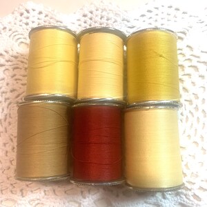 May include: A collection of seven spools of thread in various shades of yellow, brown, and red. The spools are arranged in two rows, with the thread wound around silver metal cylinders. The background is a white, crocheted doily.
