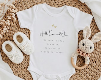 May include: White baby onesie with the text "Hallo Oma und Opa" and a sweet message. The onesie is paired with white crocheted baby shoes and a crochet bunny rattle. The items are arranged on a woven mat.