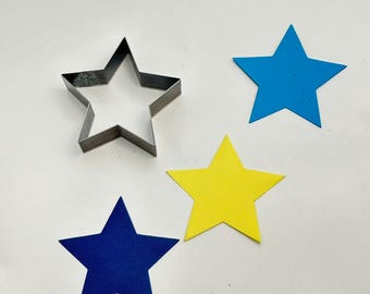 May include: A metal star-shaped cookie cutter next to three colorful star cutouts. The cutouts are in blue and yellow. The cookie cutter is made of silver metal. The stars are on a white surface.