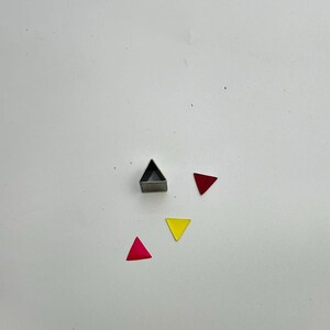 May include: A metal triangle cookie cutter next to three colorful triangle shapes. The shapes are red and yellow. The cookie cutter is silver and has a hollow triangle shape.