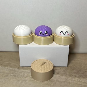 Pode incluir: Três brinquedos dim sum, cada um com um topo de cor diferente e um rosto sorridente. Os dim sum estão em pequenas cestas de cor bege. Um brinquedo dim sum adicional é mostrado separadamente.