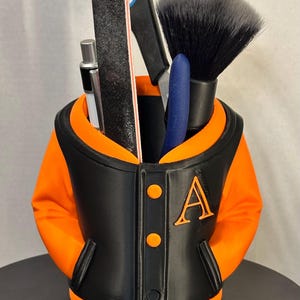 May include: A black and orange varsity jacket-shaped desk organizer holding various tools. The organizer has an orange interior and features the letter "A" on the front. Tools include scissors, a makeup brush, and a nail file.