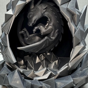 May include: A gray dragon figurine nestled inside a geometric, rocky-textured egg. The dragon is curled up, with its wings and tail wrapped around its body. The egg has a jagged, crystalline exterior, creating a fantasy-themed sculpture.