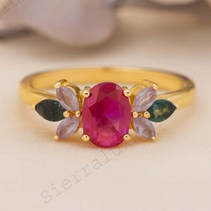 Op de afbeelding: Een gouden ring met een grote, ovaal geslepen robijn als centrale steen. De ring is versierd met kleinere edelstenen in groen, paars en lavendel tinten. Het ontwerp is bloem geïnspireerd, met de kleinere stenen rond de centrale robijn.