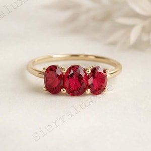 May include: A gold ring featuring three oval-cut, vibrant red gemstones. The gemstones are set closely together, creating a continuous line of color. The ring's band is a thin, polished gold, adding to its elegant design.