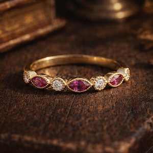 May include: A gold ring with alternating marquise-cut pink gemstones and round white diamonds. The ring is set on a dark wooden surface, with a blurred background featuring a book and other objects.