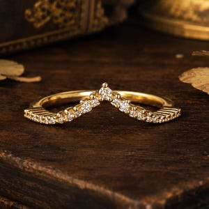 May include: A gold chevron-shaped ring featuring a series of small, clear, round-cut stones set along the band. The ring has a polished metallic finish and a pointed center design, with the stones extending across the top half of the band.