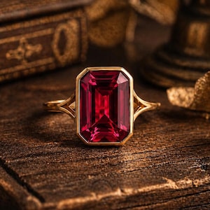 Peut inclure: Une bague en or avec une grande pierre précieuse rouge foncé de taille émeraude rectangulaire montée sur un anneau en or poli. Le sertissage présente un design à tige fendue qui se connecte à la pierre centrale, révélant une couleur vive.