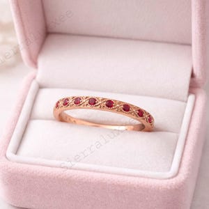 Könnte beinhalten: Roségoldener Ring mit filigranem Design, besetzt mit kleinen, runden roten Edelsteinen. Der Ring wird in einer rosa Samt-Ringbox präsentiert, die seine filigranen Details und seinen eleganten Stil hervorhebt.