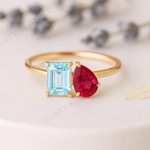 May include: A gold ring featuring two gemstones: a rectangular light blue aquamarine and a teardrop-shaped red ruby. The ring's band is a thin, polished gold.
