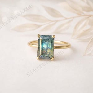 Puede incluir: Un anillo de oro con una piedra preciosa rectangular multicolor. La piedra preciosa tiene tonos de azul, verde y amarillo. La banda del anillo es de oro fino y pulido. El engaste tiene cuatro garras.