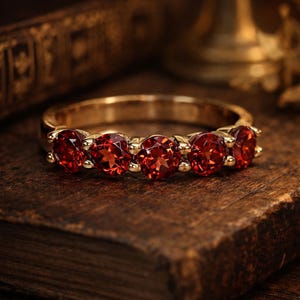 May include: A gold band ring featuring five round, vibrant red gemstones. The ring is set on a wooden surface, with a blurred background. The gemstones are evenly spaced and appear to be faceted.
