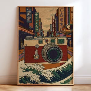 May include: A framed art print featuring a vintage camera design with a Japanese city backdrop and ocean waves. The camera is red and cream-colored, with a large lens. The background includes buildings and Japanese text.