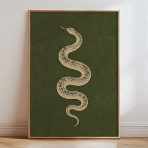 May include: A framed wall art print featuring a beige, textured snake illustration with a forked tongue, positioned in a vertical S-curve against a solid, deep olive green background. The frame is made of light-coloured wood with a thin profile.