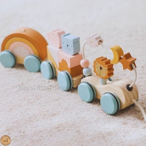 Puede incluir: Tren de juguete de madera para tirar con bloques de colores pastel, un arcoíris y un coche con temática de león. El tren tiene ruedas azul claro y una cuerda para tirar. Los bloques tienen diseños de oso y nube.