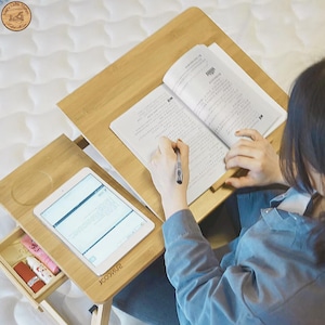 May include: A bamboo laptop desk with a tablet and a book. The desk has a small drawer with beauty products. The tablet displays text. The book is open, and a pen is held over the pages. The desk is on a bed.