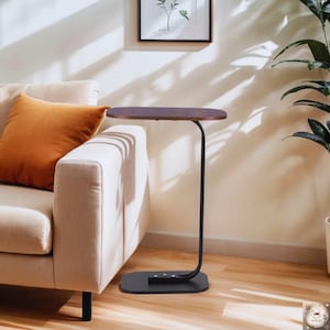 May include: A contemporary side table featuring a dark brown, oval tabletop and a black, curved metal frame. The table has a rectangular base. It is placed beside a beige sofa with an orange cushion. The background includes a plant and a framed picture.