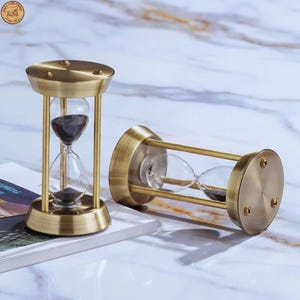 Op de afbeelding: Twee zandlopers in antieke stijl met messingkleurige metalen frames. De glazen bollen bevatten donker zand. Eén zandloper staat rechtop, terwijl de andere op zijn kant ligt. De zandlopers staan op een wit en grijs marmerpatroon oppervlak.