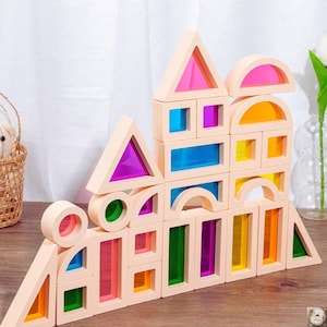 May include: A collection of wooden building blocks in various shapes, including triangles, rectangles, and arches. The blocks have transparent coloured inserts in shades of pink, purple, blue, green, and yellow. The blocks are arranged in a stacked formation.