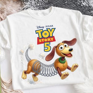 Puede incluir: Una camiseta blanca con un gr&aacute;fico del personaje Slinky Dog de Toy Story. Sobre el personaje, el texto dice Disney Pixar Toy Story 5. El perro tiene un cuerpo marr&oacute;n, una secci&oacute;n media de resorte met&aacute;lico y un collar verde con una placa dorada.