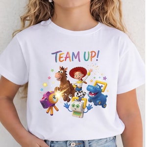 May include: A white short-sleeve t-shirt featuring a colorful graphic print with the text "TEAM UP!" above cartoon characters including a brown horse, a cowgirl, a blue hippo, a camera, and a small robot, surrounded by small multi-colored stars.