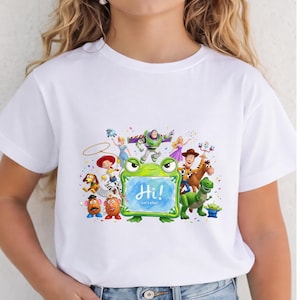 May include: A white short-sleeved t-shirt featuring a colorful graphic print of various Toy Story characters surrounding a green frog-shaped frame. The center of the frame contains a blue watercolor background with the white text Hi! Let's play!