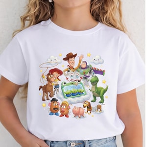 May include: A white short-sleeved t-shirt featuring a colorful graphic print of various animated characters including a cowboy, a space ranger, a cowgirl, a horse, a dinosaur, a dog, a pig, and several small green aliens in a blue container.