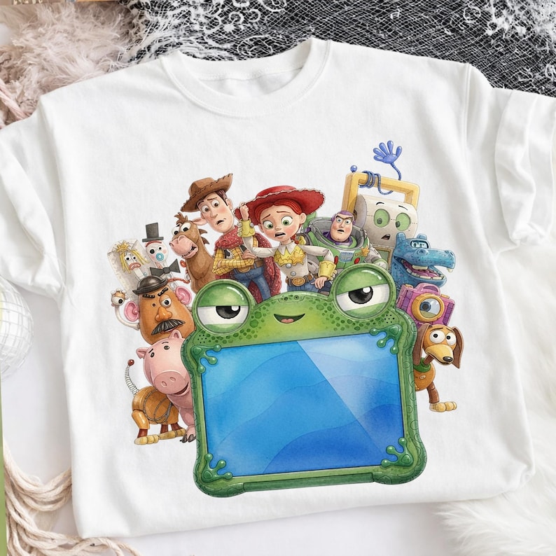 Puede incluir: Una camiseta blanca de manga corta con un estampado gr&aacute;fico de varios personajes animados de Toy Story que rodean un marco grande de color verde con forma de rana y centro azul. Los personajes incluyen a Woody, Jessie, Buzz Lightyear, Sr. Patata, Hamm, Rex y Slinky Dog.