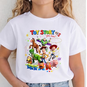 May include: A white short-sleeved t-shirt featuring a colorful graphic print of Toy Story characters including Woody, Buzz Lightyear, Jessie, Rex, Bullseye, Slinky Dog, Hamm, Mr. and Mrs. Potato Head, and the Aliens, with the text Toy Story yee haw.