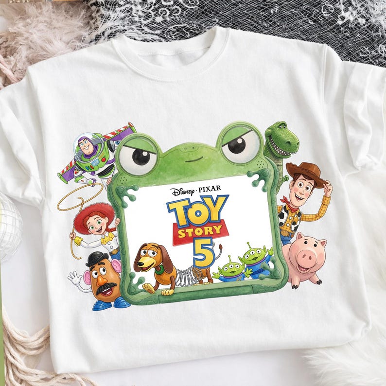 Puede incluir: Una camiseta blanca de manga corta con un estampado gr&aacute;fico de los personajes de Disney Pixar Toy Story, incluyendo a Buzz Lightyear, Woody, Jessie, Mr. Potato Head, Slinky Dog, Rex, Hamm y Aliens rodeando un marco verde con forma de rana.