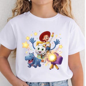 May include: A white t-shirt featuring a colorful graphic print of a red-haired cowgirl character, a blue hippopotamus, a white calculator-like character, and a purple camera on a tripod, all surrounded by yellow stars and confetti.