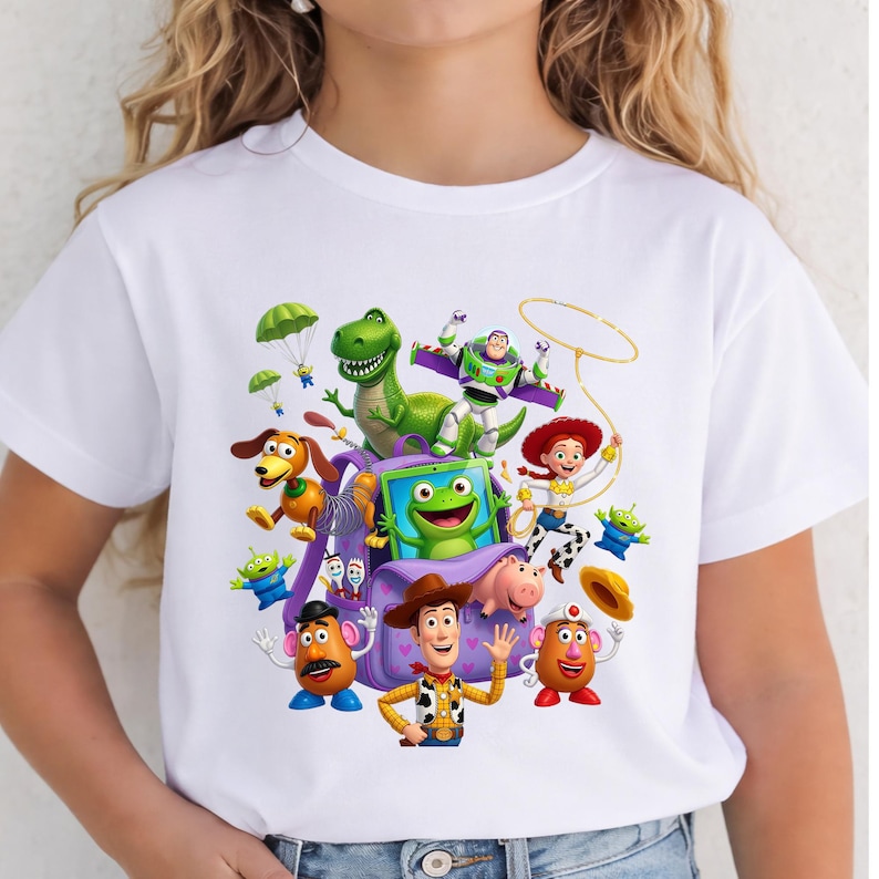 May include: A white short-sleeved t-shirt featuring a colorful graphic print of various animated characters from the Toy Story franchise, including Woody, Buzz Lightyear, Jessie, Rex, Slinky Dog, Mr. and Mrs. Potato Head, and a green frog.