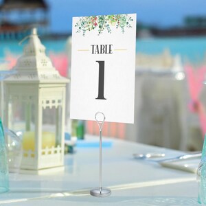 May include: A white table number card with the word "TABLE" and the number "1" in black. The card is decorated with a floral design and held by a silver metal stand. The background is a blurred outdoor scene.