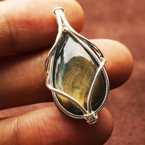 May include: A teardrop-shaped pendant featuring a polished, iridescent gemstone encased in a silver wire wrap. The stone displays shades of green, gold, and blue. The wire is intricately woven around the stone, creating a decorative frame.