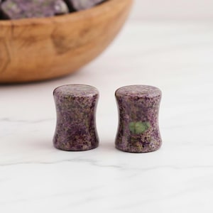 May include: Two purple and green stone plugs with a flared hourglass shape. The plugs are made of a speckled stone material and sit on a white surface. A wooden bowl filled with similar stones is in the background.