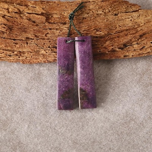 May include: Two rectangular purple stone pendants with a speckled texture, connected by a green string. The pendants are displayed against a textured wooden background and a neutral gray surface.