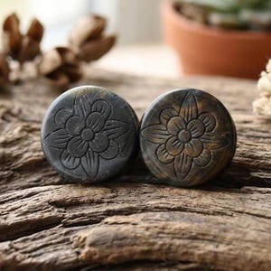 Puede incluir: Dos tapones redondos de color gris oscuro con un diseño floral tallado. Los tapones están hechos de un material similar a la piedra y descansan sobre una superficie de madera envejecida. El patrón floral es detallado y añade un elemento decorativo.