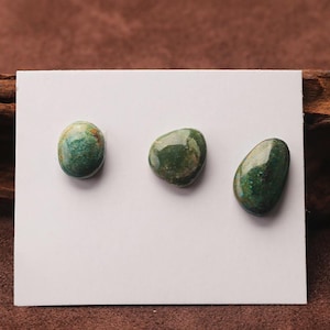 May include: Three polished, oval-shaped stones in shades of green and teal are displayed on a white card. The stones have a smooth, glossy texture and varying patterns. The background is a mix of brown and white.