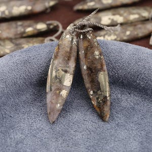 May include: Two elongated, teardrop-shaped pendants with a natural, earthy appearance. The pendants are primarily brown and gray with shimmering, metallic gold flecks. A gray string is attached at the top, suggesting they are designed to be worn as jewelry.
