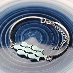 May include: Silver-toned bracelet featuring a cluster of six light blue fish charms. The bracelet has a simple, elegant design with a delicate chain for adjustable sizing. The fish charms are detailed with black outlines, adding contrast to the light blue enamel.