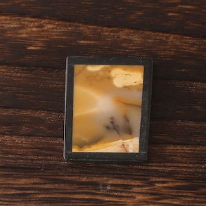 May include: A rectangular pendant with a black frame. The center features a natural stone with a mix of beige, cream, and brown hues. The stone has a unique, organic pattern. The pendant is set against a dark wooden surface.