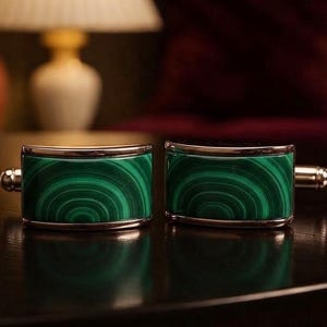 以下が含まれることがあります： 光沢のあるシルバー調の金属フレームと、独特の同心円状の縞模様を持つ緑色のマラカイト石のインレイが特徴の長方形のカフスボタン一対。天然石の質感を強調するように、2つが並べて配置されています。