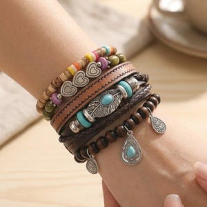 Puede incluir: Una pila de pulseras bohemias en varios estilos. Las pulseras incluyen cuentas de madera, bandas de cuero, detalles turquesa y dijes plateados. Una pulsera presenta un diseño de pluma, otras tienen colgantes de corazón y lágrima.